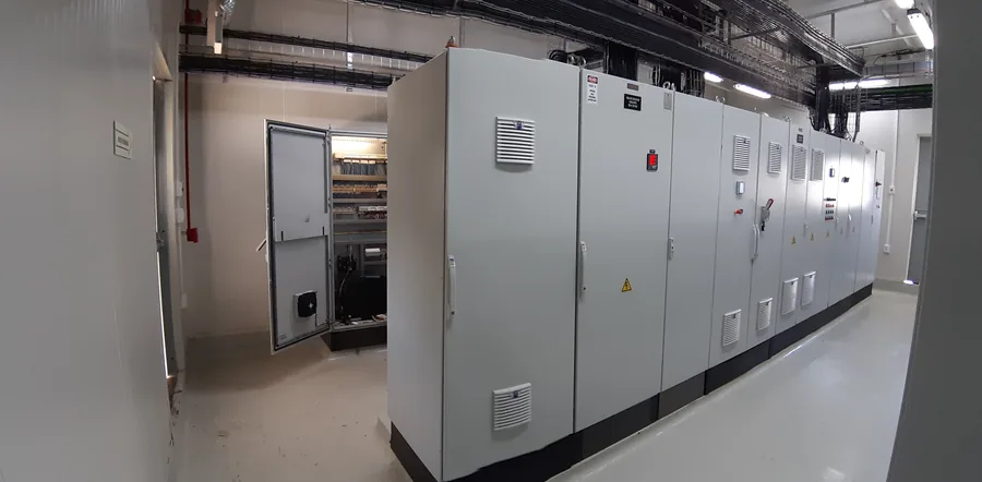 Cuarto eléctrico con una fila de tableros de control y fuerza montados contra la pared; uno de ellos abierto durante el cableado