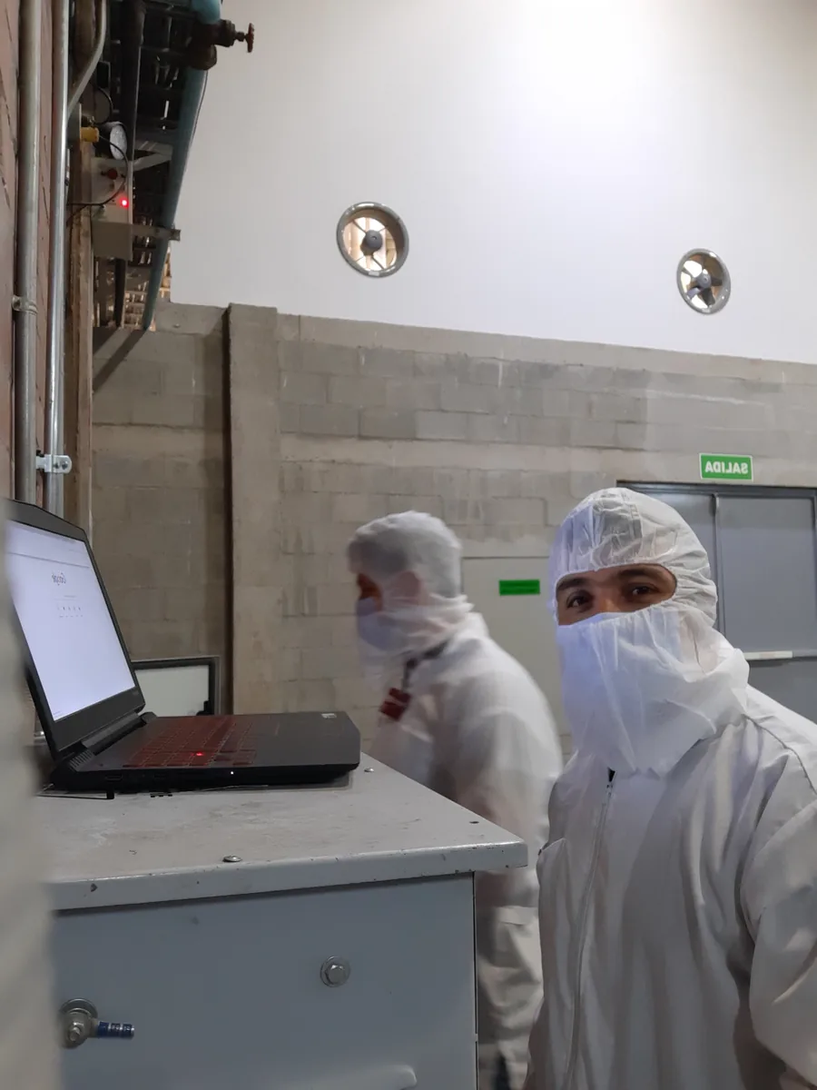Dos técnicos con traje y cofia de planta revisan el programa en un portátil apoyado sobre el tablero, dentro de una sala de producción
