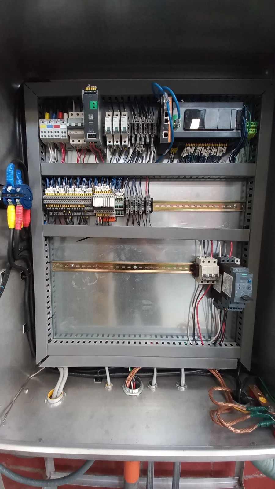 Tablero en acero inoxidable con PLC compacto, switch de red y borneras; guantes dieléctricos colgados en la puerta