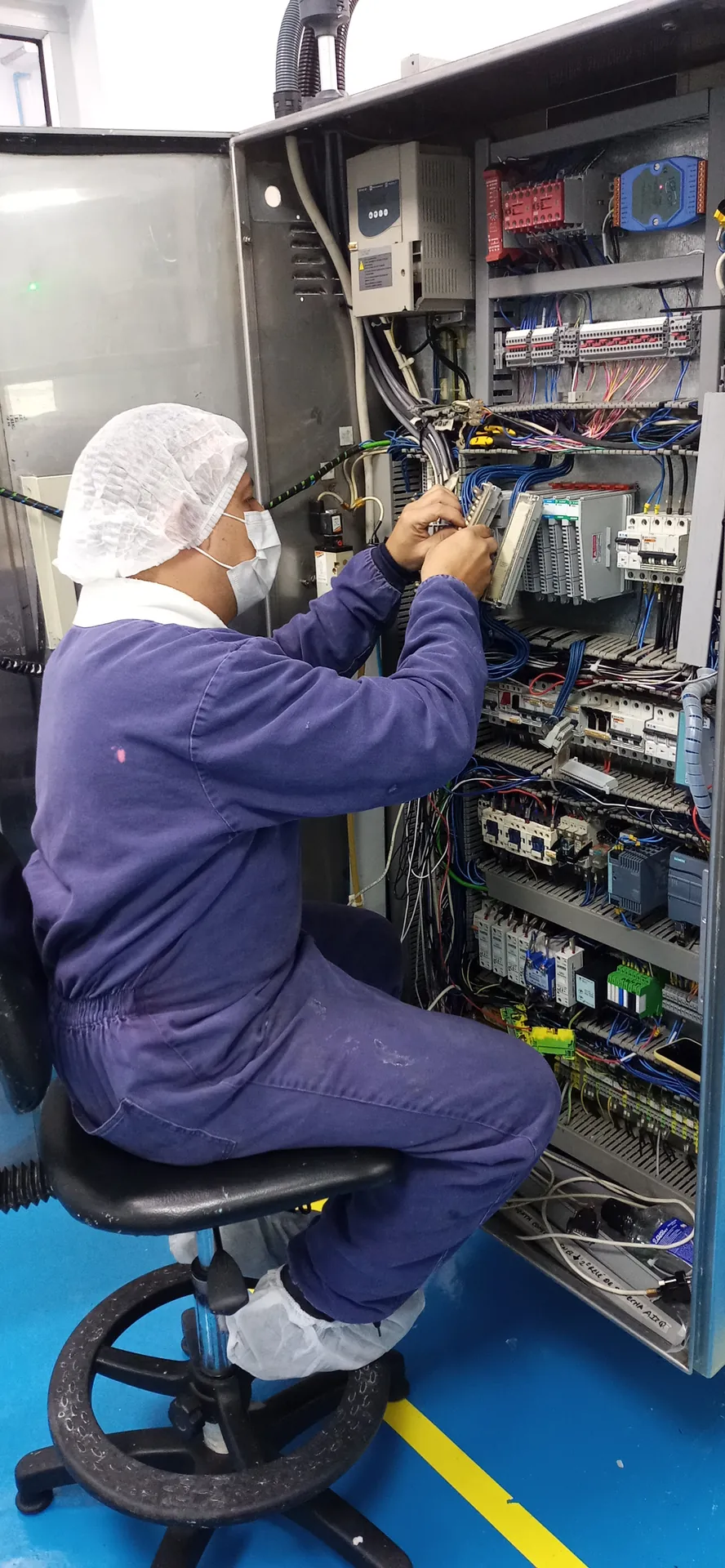 Técnico con overol y cofia trabaja en el cableado interno de un tablero de control dentro de una planta de alimentos